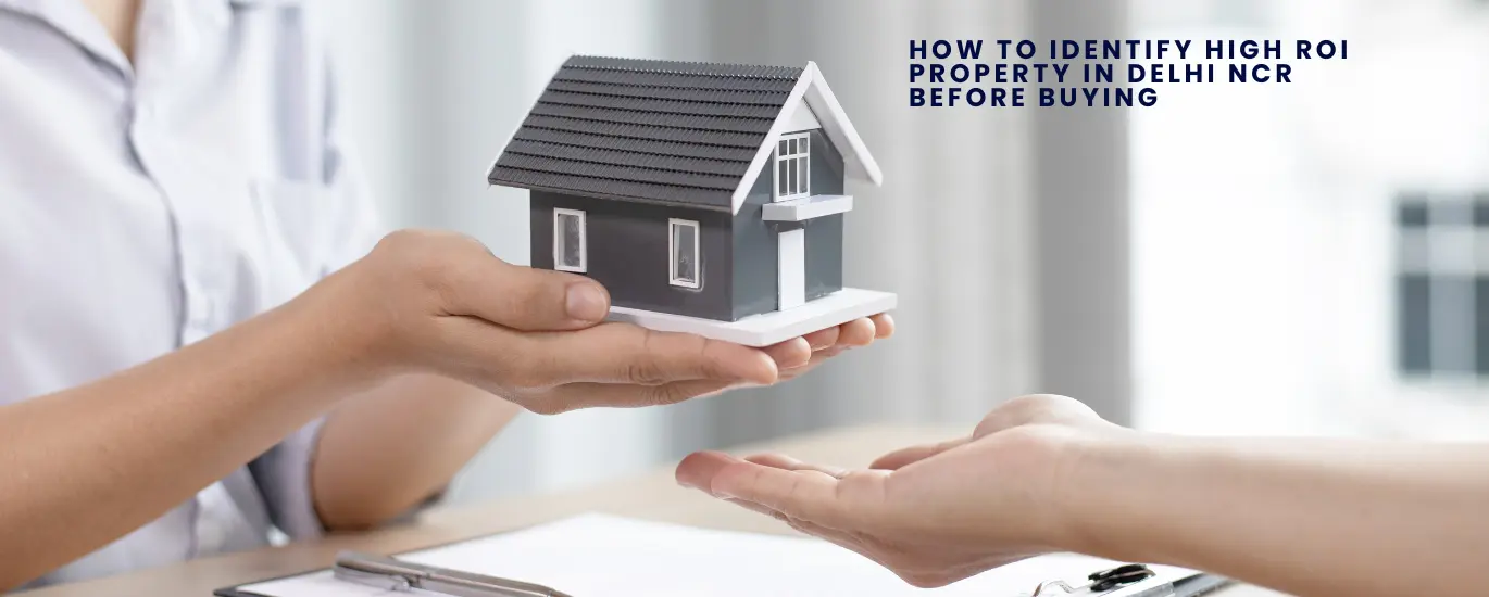 A person hands over a small model of a house to another person, symbolizing a real estate transaction, with the text "How to Identify High ROI Property in Delhi NCR Before Buying" in the background.